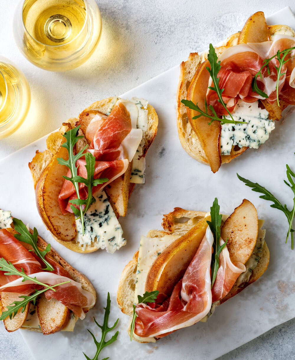 Flat lay of crostini appetizers with blue cheese, caramelized pears, prosciutto, and arugula, catered by Joel Catering, New Orleans' elite catering service