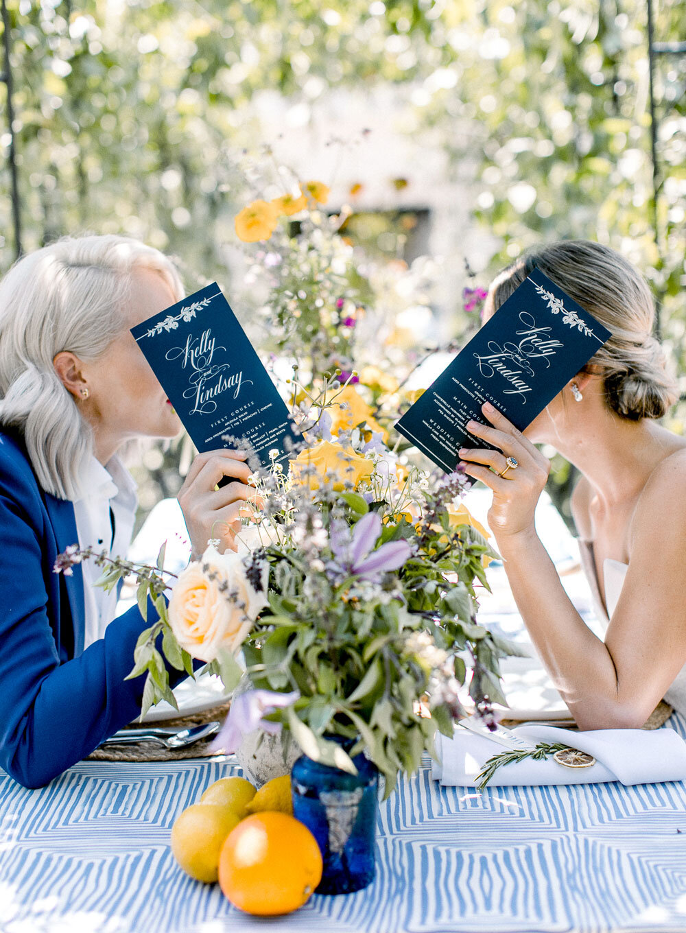 Two brides reading their wedding invitations at an outdoor table adorned with yellow, purple, and white flower arrangements and a blue patterned runner, captured by Joel Catering, New Orleans' LGBTQ-friendly event specialist