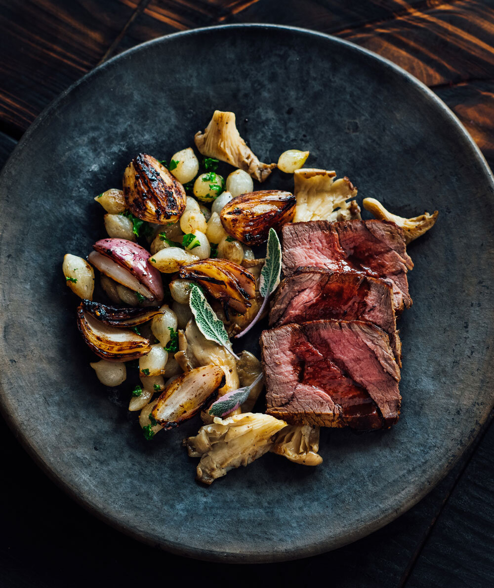 Sliced juicy filet mignon topped with a rich reduction sauce, accompanied by caramelized oyster mushrooms and pearl onions, garnished with white sage, a gourmet entrée by Joel Catering in New Orleans.